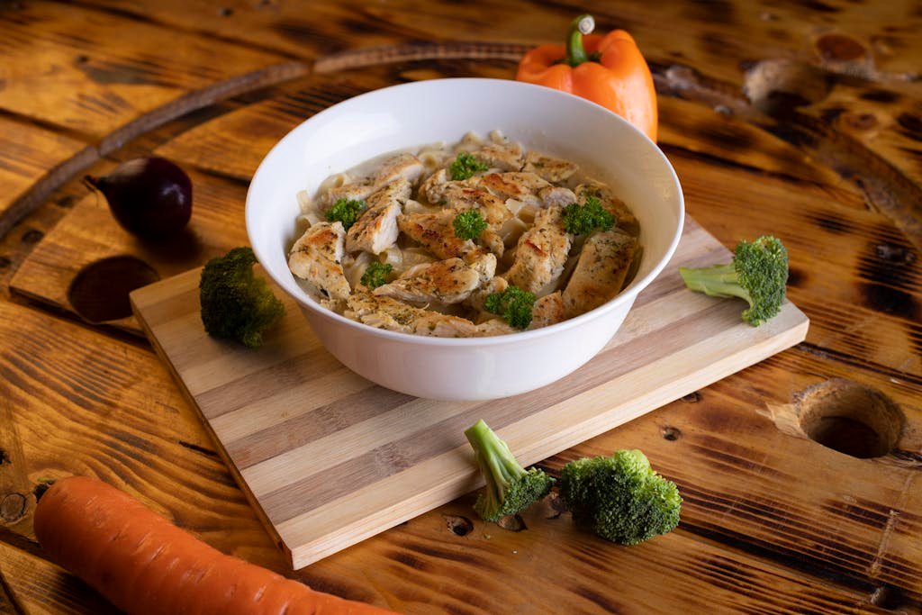 Close-up of chicken pasta in a white bowl with fresh vegetables around on a rustic table.