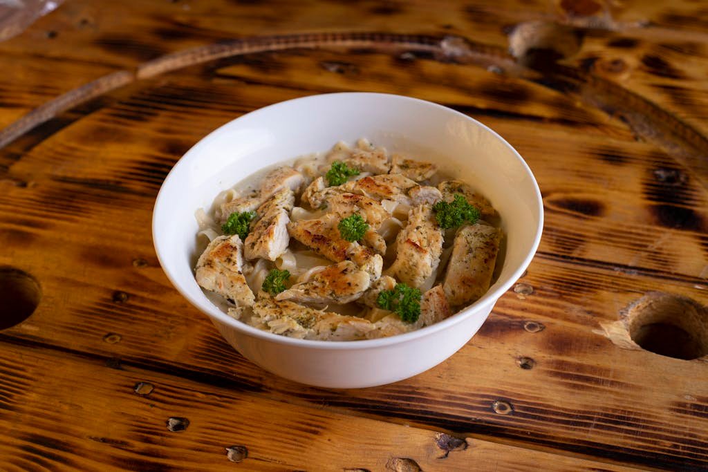 Tasty chicken pasta with herbs in a white bowl on rustic wooden table.